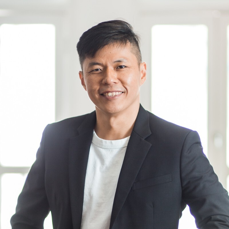 Smiling man wearing a red sweater and checkered shirt standing outdoors with blurred trees in the background.