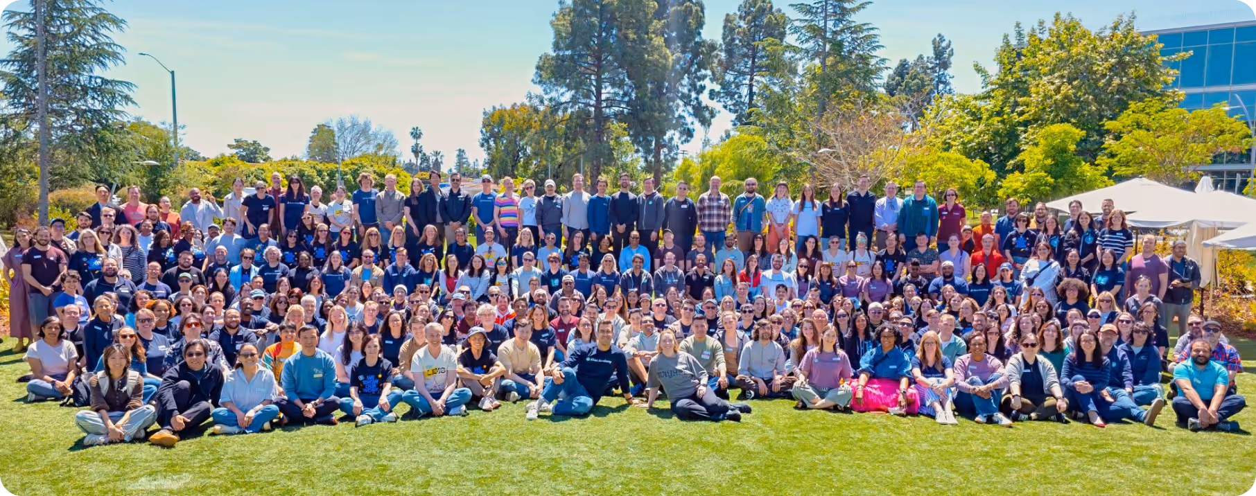 Large diverse group of people posing on grass outdoors with trees and buildings in the background.