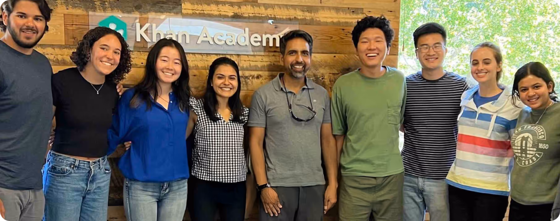 Diverse group of eight smiling people standing in front of a wooden wall with Khan Academy sign.