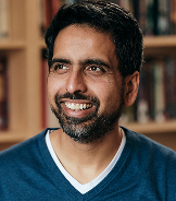 Smiling man with dark hair and beard wearing a blue sweater over a white shirt in a book-filled room.