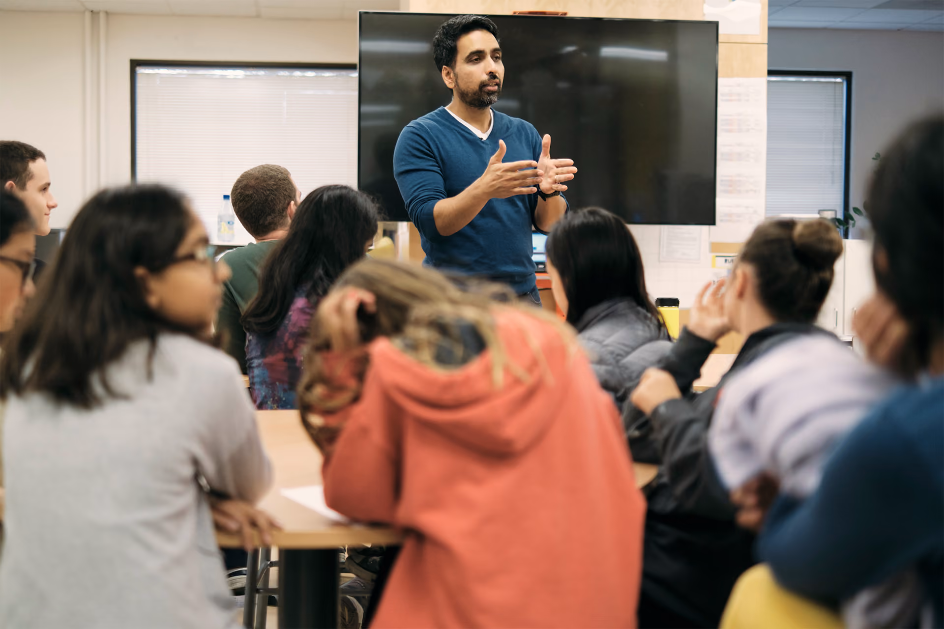 A man in a blue sweater gestures while speaking to a classroom of attentive students.