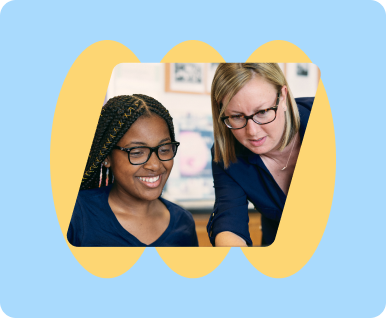 Teacher with glasses smiling with another female learner while looking at something on a screen in a classroom.