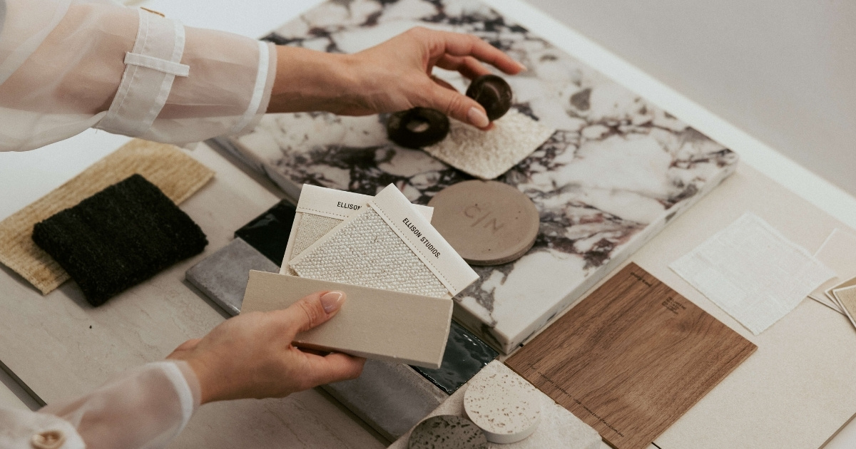 Hands holding and arranging various fabric swatches and material samples on a marble surface.