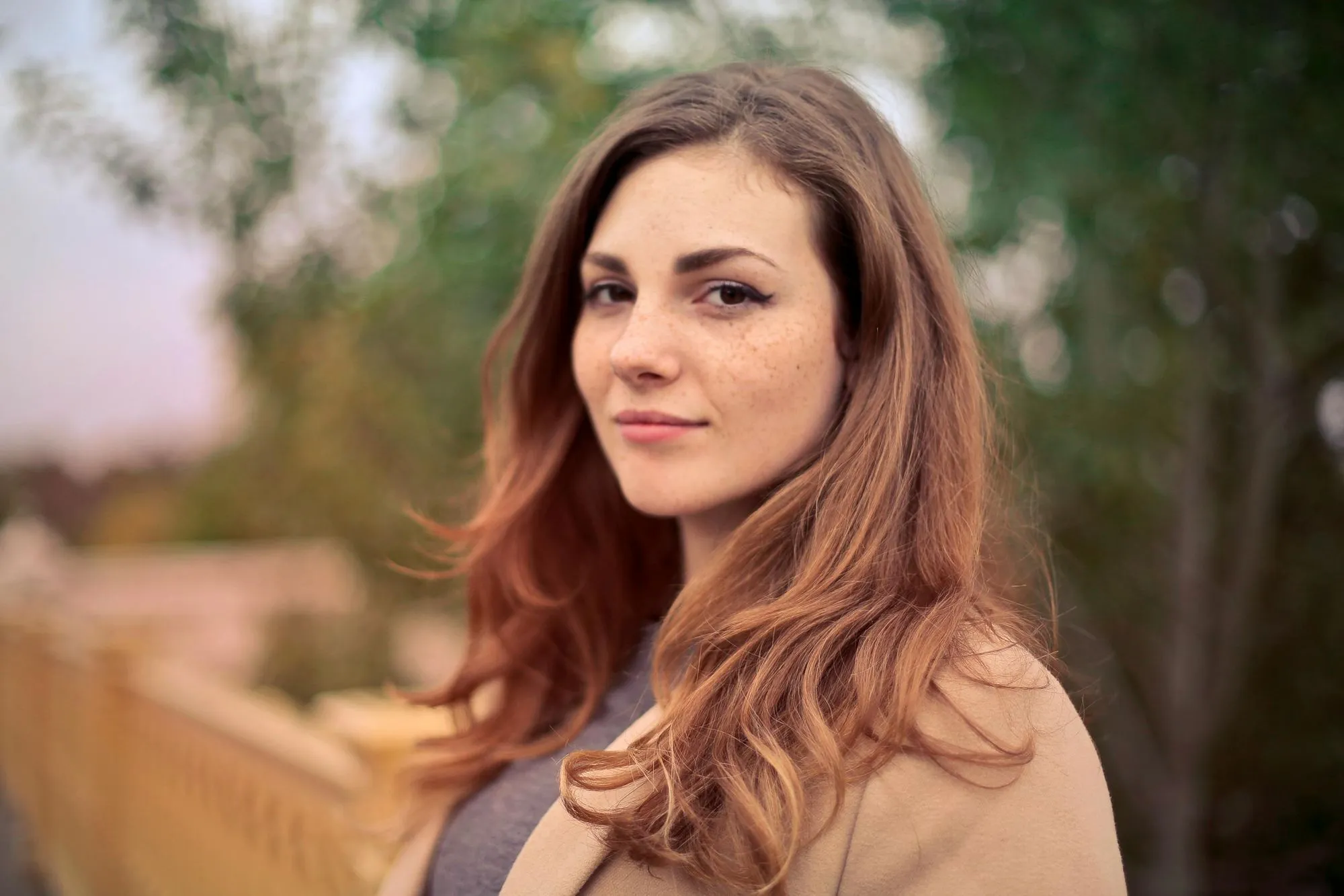 Young woman with long reddish-brown hair and freckles wearing a beige coat outdoors with blurred greenery in the background.