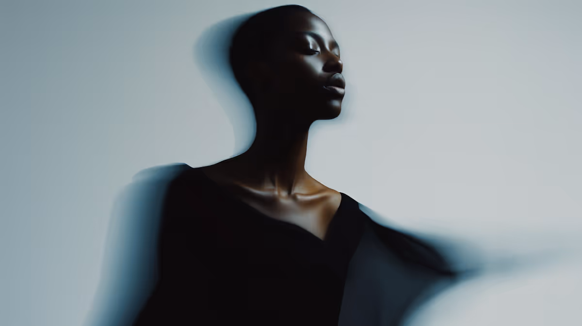 Portrait of a dark-skinned woman with closed eyes in a black outfit and blurred motion effect on a light gray background.