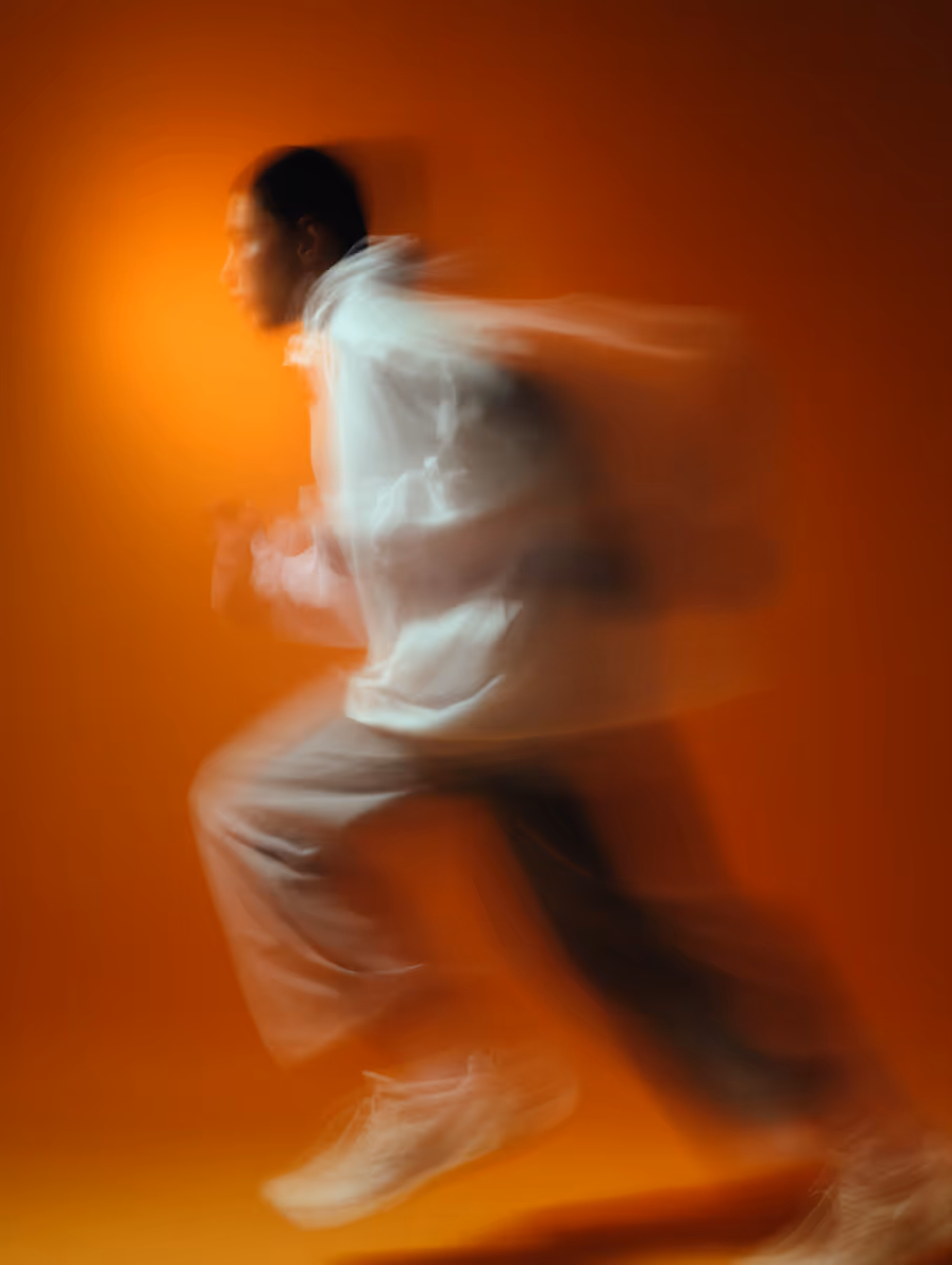 Blurred motion of a person running against an orange background wearing white sneakers and beige pants.