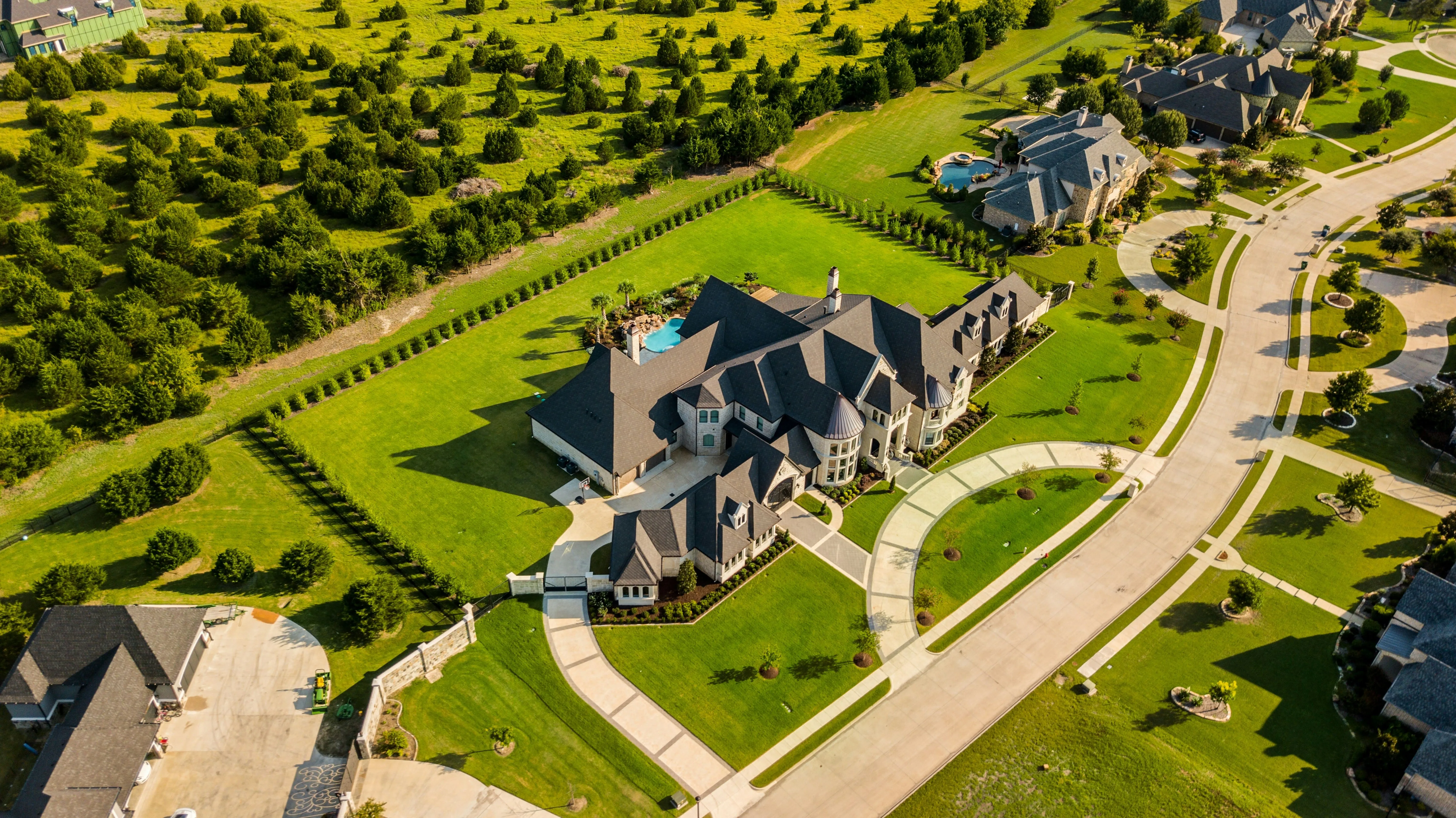 Aerial view of a large suburban house with a dark grey roof, surrounded by a green lawn and curved concrete driveways.