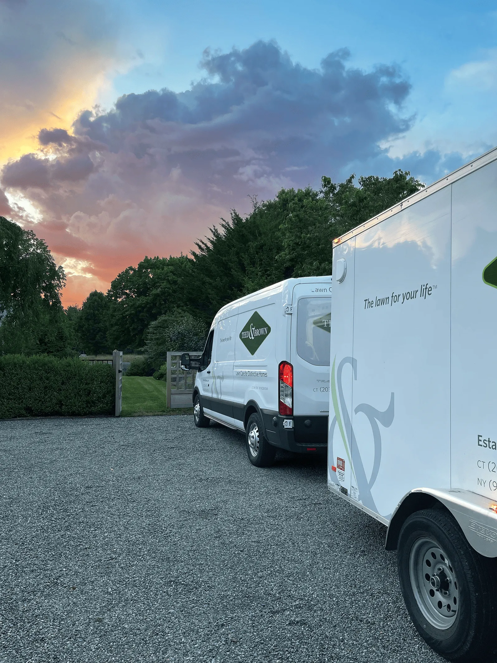 Two white vehicles with green company logo parked on gravel near a green gate with trees and a colorful sunset sky in the background.
