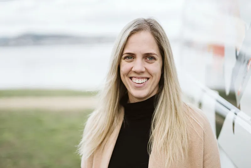 Smiling woman with long blonde hair in black top and beige coat outdoors