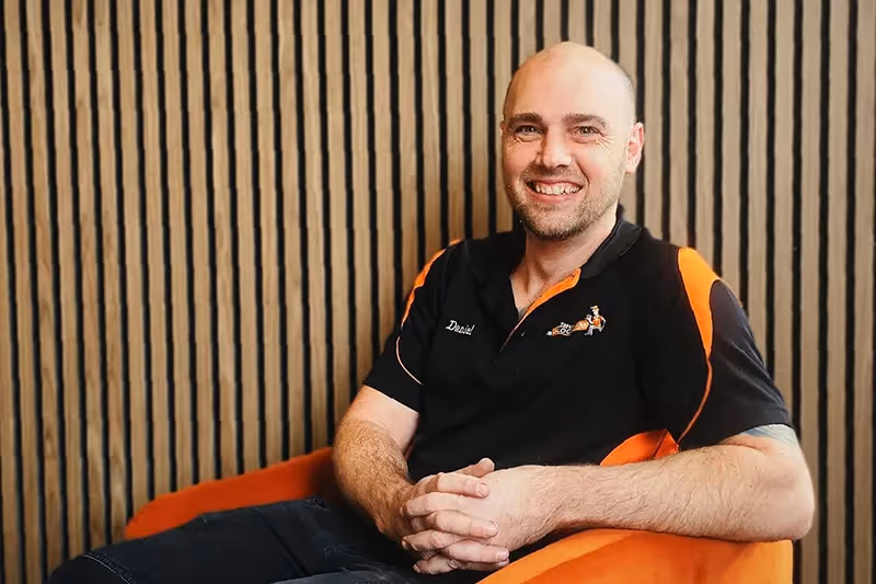 Smiling professional in black and orange polo shirt sitting against striped wall