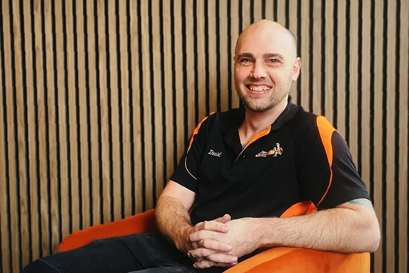 Smiling professional in black and orange polo shirt sitting against striped wall