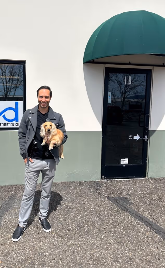 A person stands outdoors holding a small brown dog, framed by a building with a green awning and a reflective glass door. "We're never doing this again..." one mutters, glancing at the pup playfully wagging its tail.