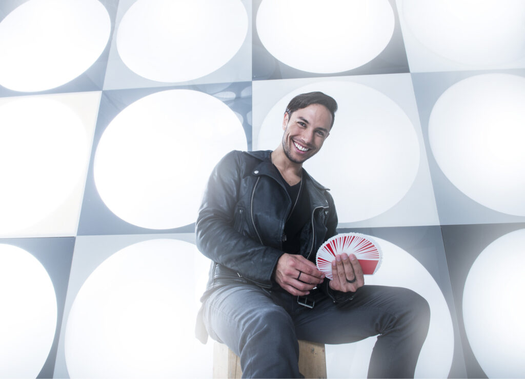 Magician Alex Ramon smiling while holding a fan of playing cards in a bright studio setting.