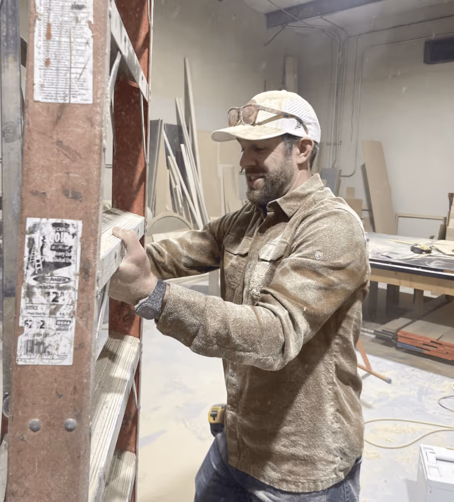 Houston wearing a cap and work jacket, handling a ladder in a dusty woodworking shop.