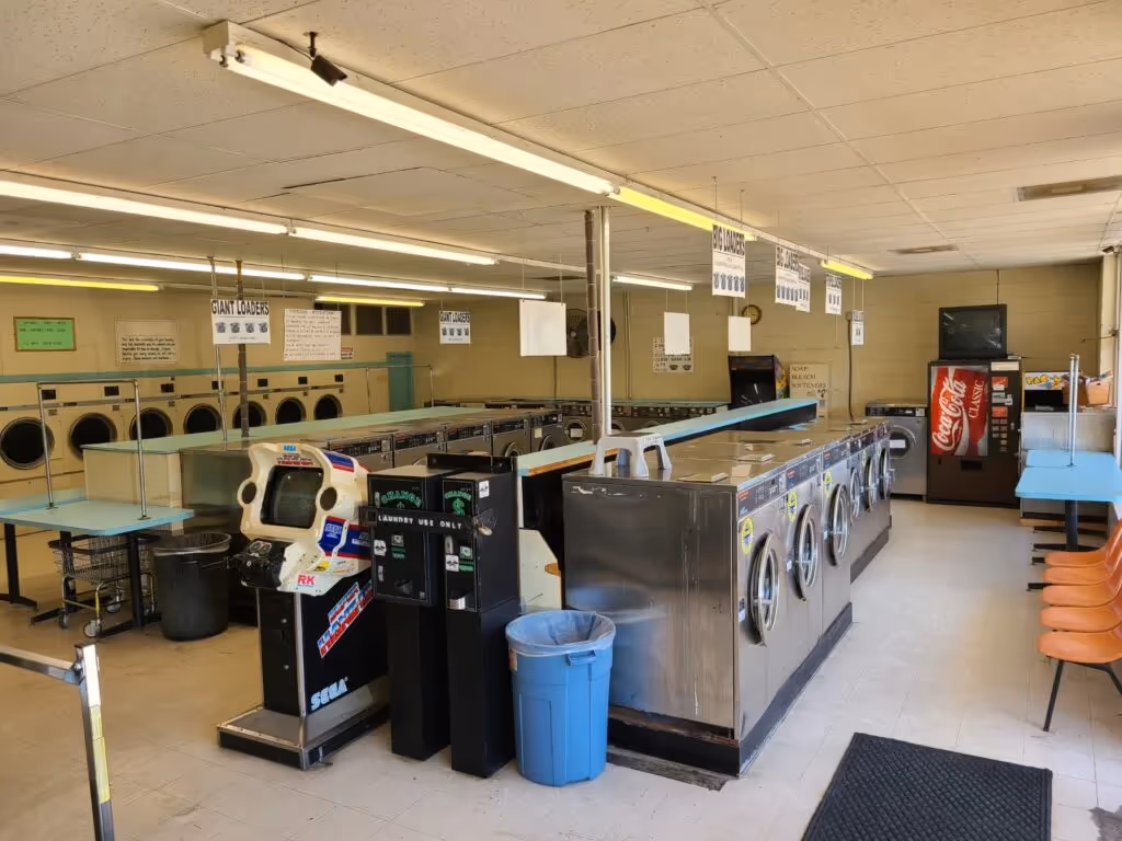 A clean and organized laundromat with large washing machines, a Coca-Cola vending machine, and spacious seating areas.