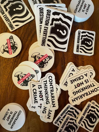 A pile of black and white stickers spread across a wooden surface. Designs include slogans like “Contrarian Thinking Is Not a Crime” and “Question Everything.”