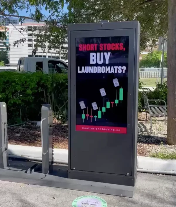 Digital billboard in a public outdoor setting displaying the text "SHORT STOCKS, BUY LAUNDROMATS?" with a candlestick chart and rising laundromat icons. Branding at the bottom reads "ContrarianThinking.co".