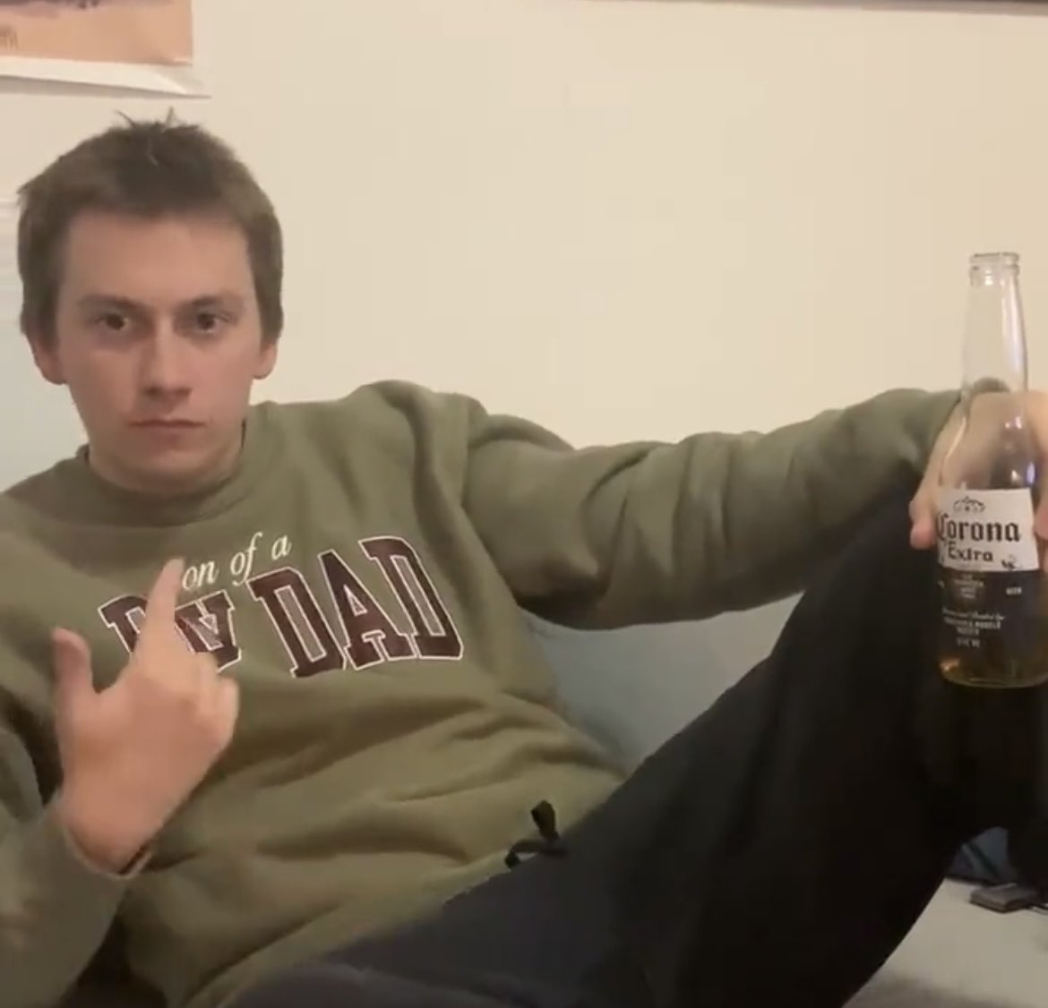 Man sitting casually on a couch wearing a sweatshirt that says “Son of a Day Dad,” pointing at himself with one hand and holding a Corona Extra beer in the other.