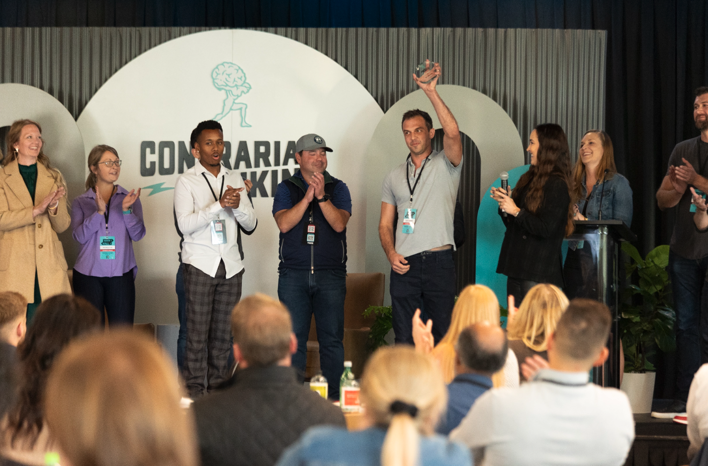 Photo of a live Contrarian Thinking event. A man is raising a trophy while others on stage clap, with audience members watching from below.