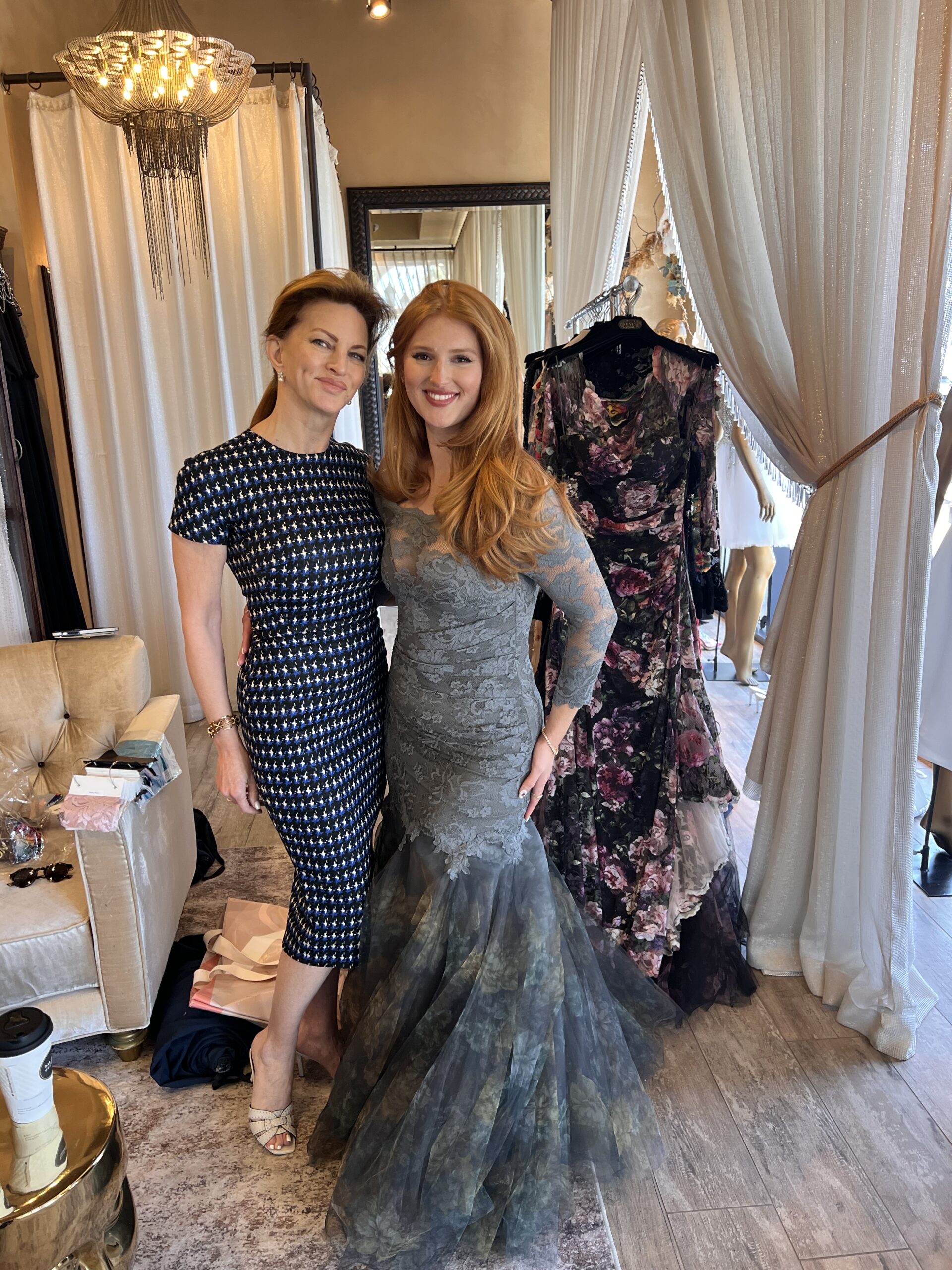 Two women posing together in a boutique, one in a fitted gown, surrounded by elegant dresses.