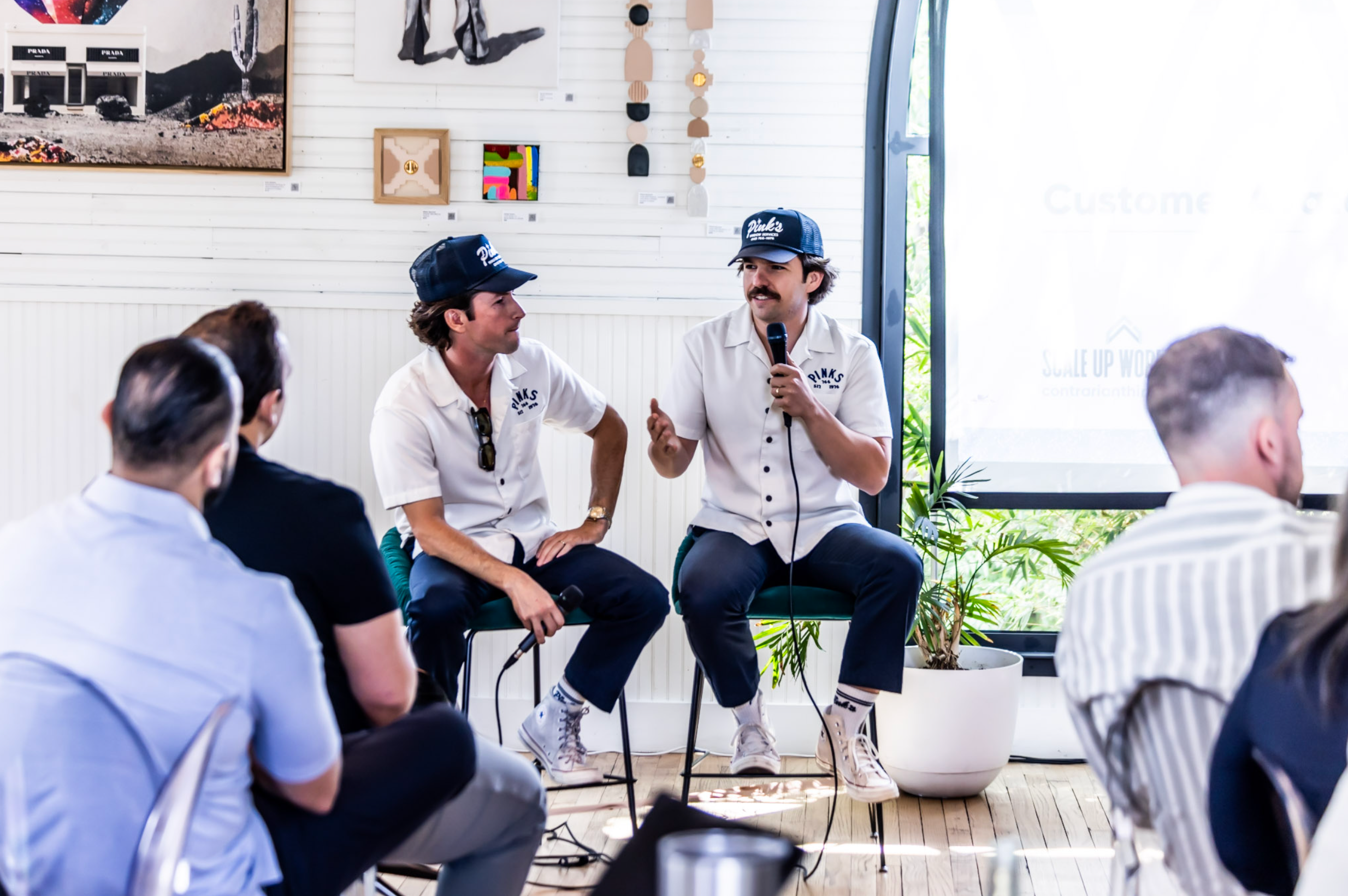 Two men in matching shirts and caps speaking at a business event in front of an audience, sharing their entrepreneurial story.