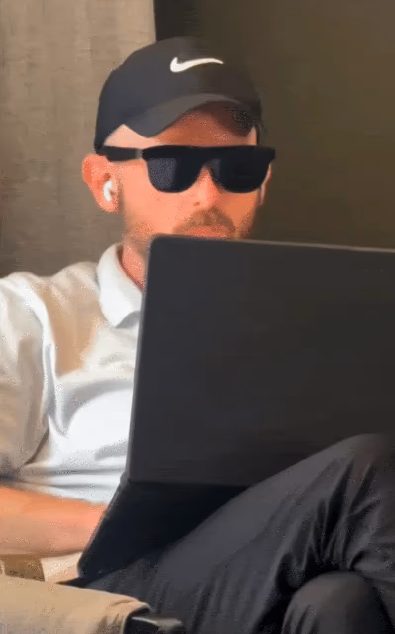 Man wearing a black Nike cap, sunglasses, and white earbuds sitting with a laptop on his lap, focused on the screen.