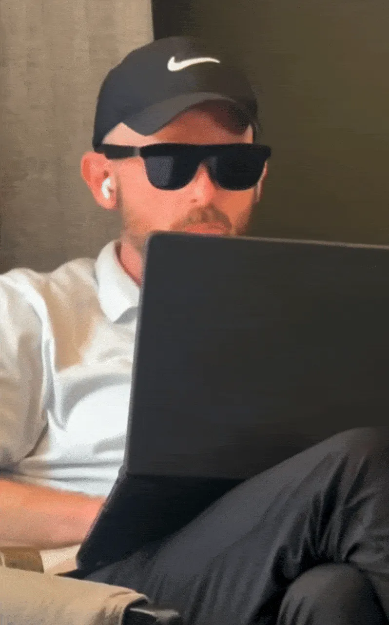 Man wearing a black Nike cap, sunglasses, and white earbuds sitting with a laptop on his lap, focused on the screen.