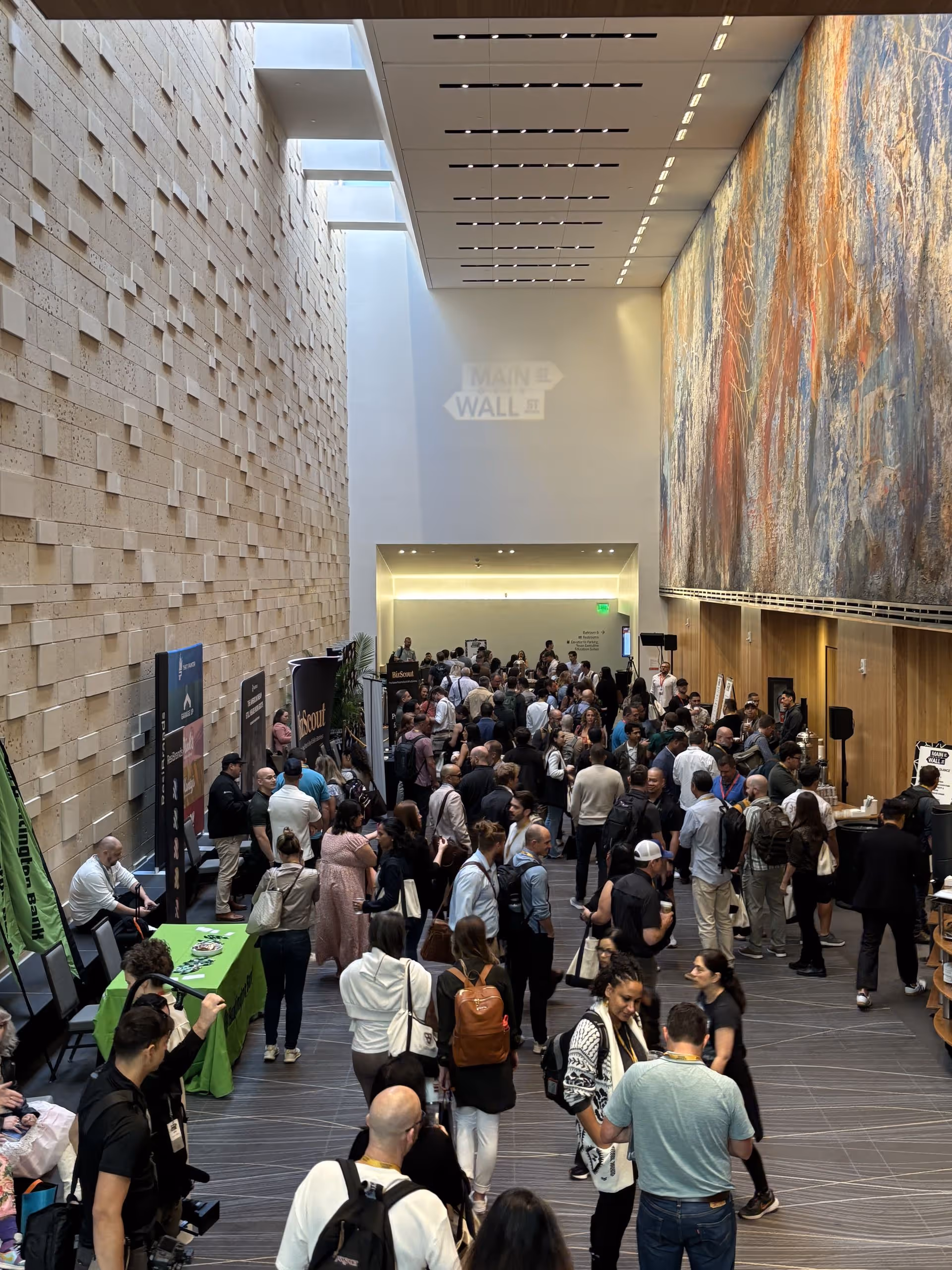 Crowded networking area at Main Street Over Wall Street, with attendees talking, visiting vendor tables, and moving through a bright atrium with tall textured walls and large mural artwork.