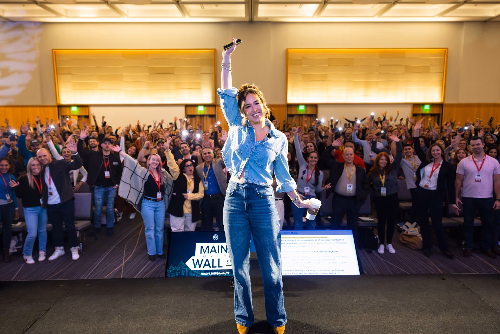 Codie Sanchez stands on stage holding a microphone at Main Street Over Wall Street, smiling as a large, enthusiastic audience cheers behind her.