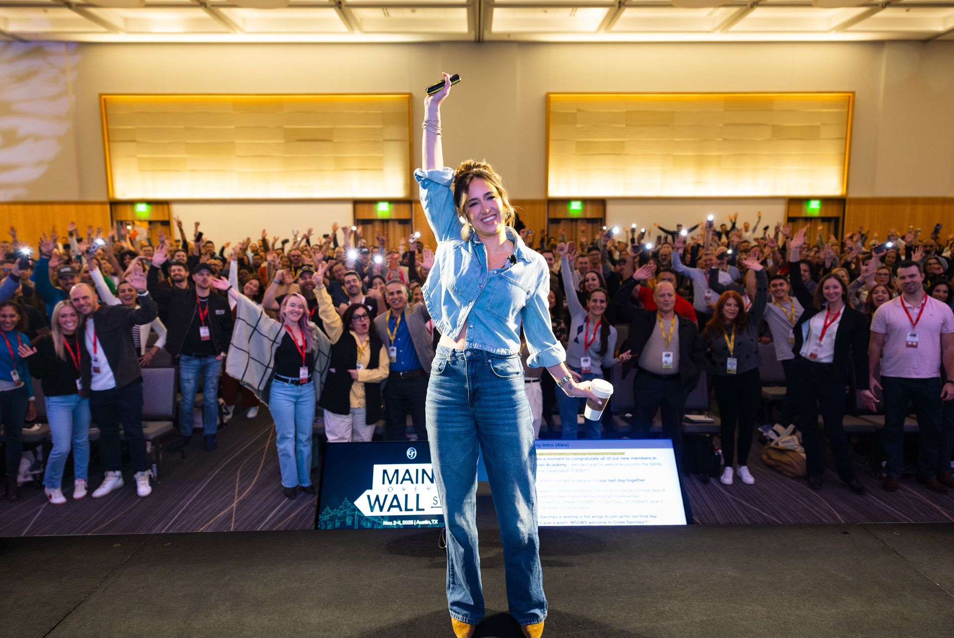 Codie Sanchez stands on stage holding a microphone at Main Street Over Wall Street, smiling as a large, enthusiastic audience cheers behind her.