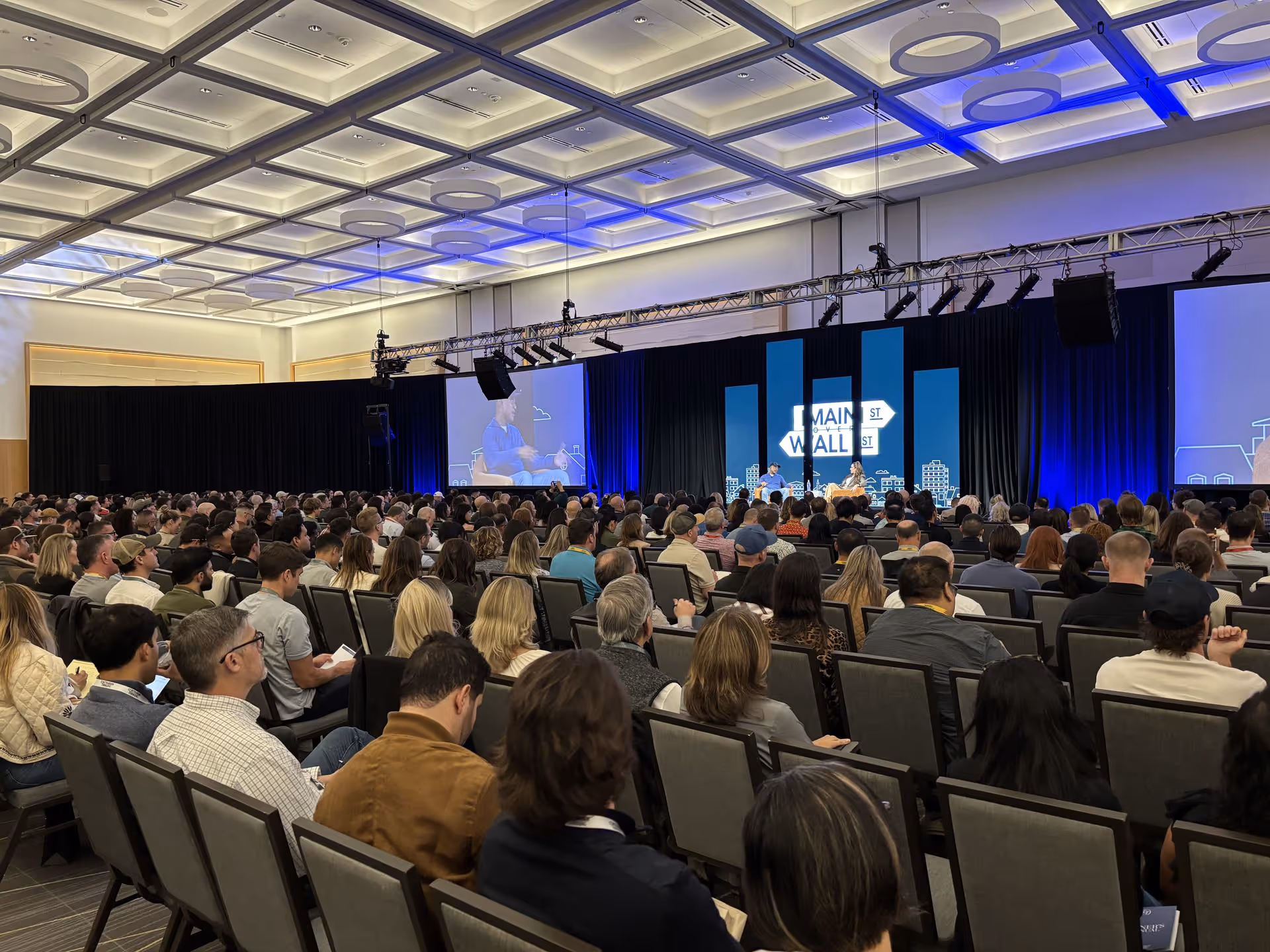 A full auditorium of attendees seated and listening to a live panel on stage at Main Street Over Wall Street, with large screens and blue stage lighting.