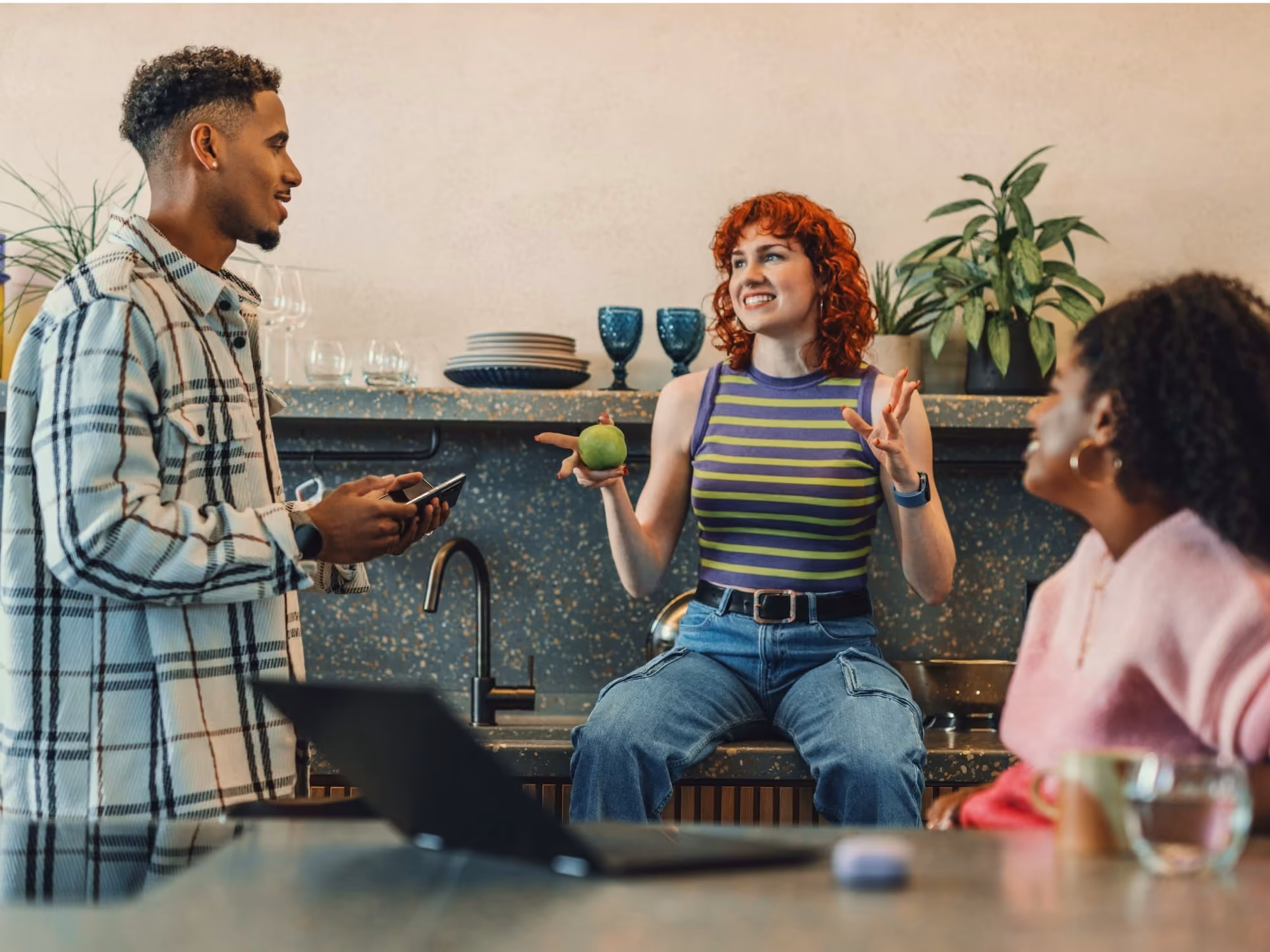 Three diverse young adults casually talking in a modern kitchen, one standing with a phone, one sitting on the counter holding a green apple, and one seated nearby, with plants and kitchenware in the background.