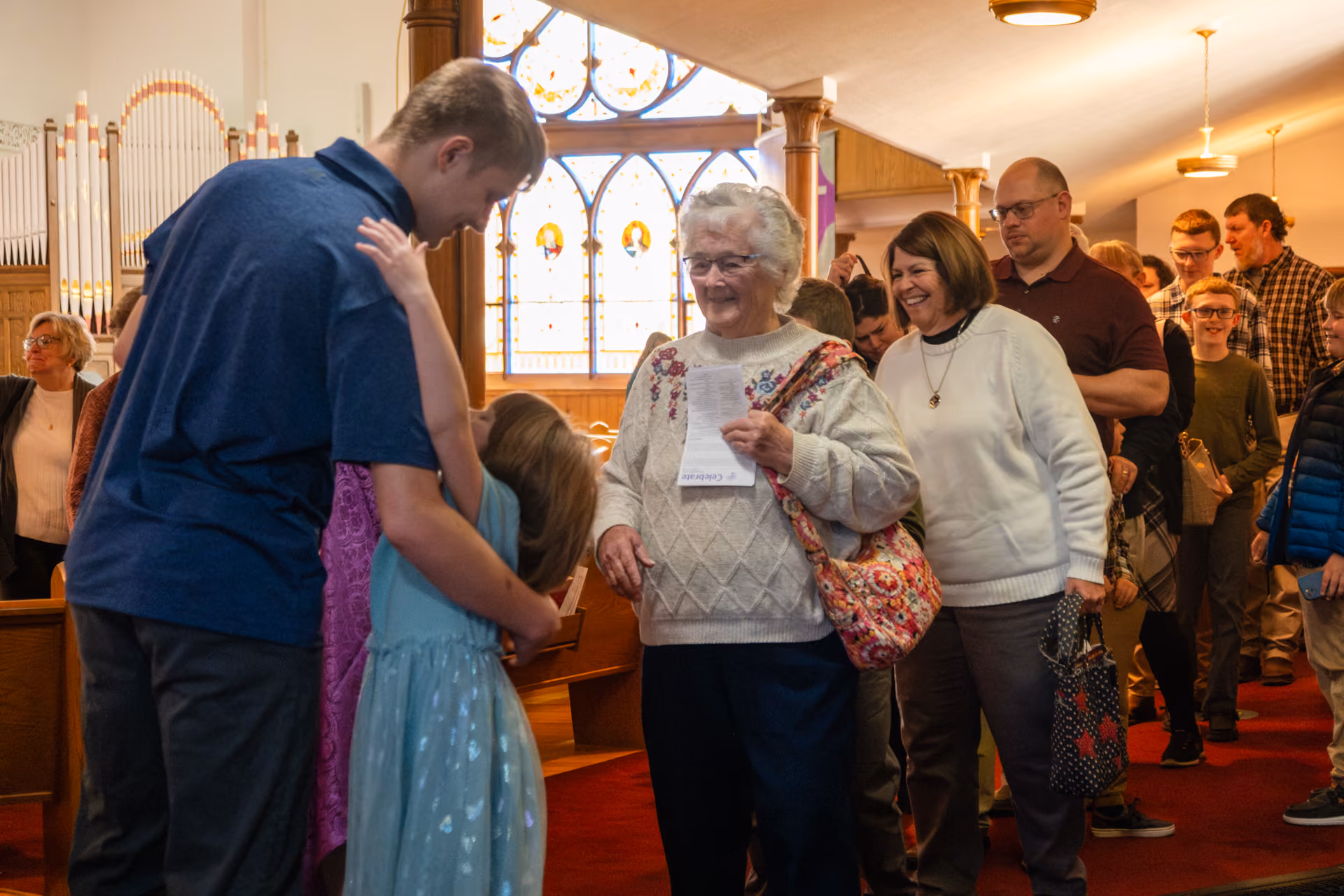 People inside a church warmly greeting and hugging near stained-glass windows and organ pipes, with an elderly woman smiling in the center holding a paper and others standing in a line behind her.