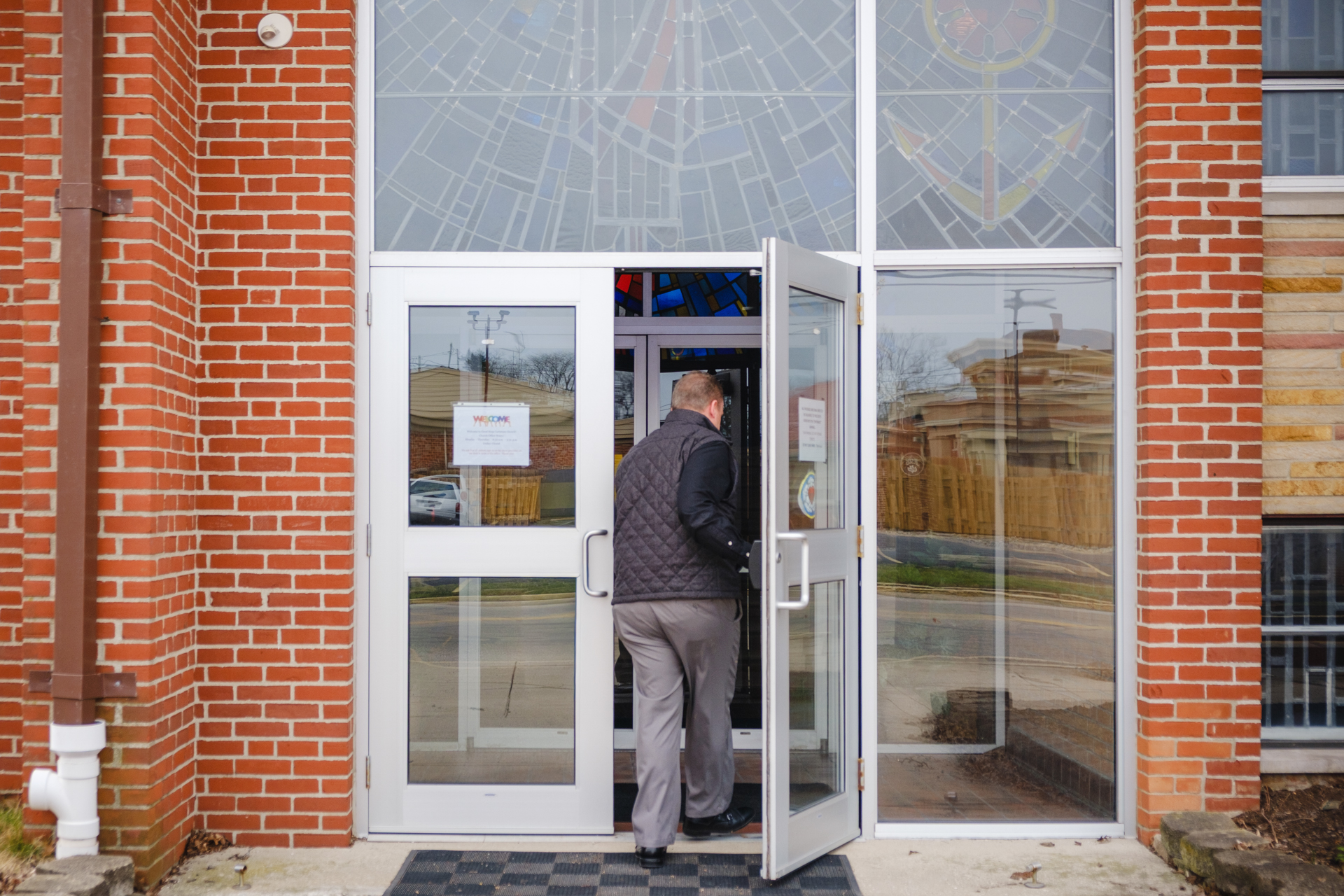 Man wearing a black quilted vest entering the church building through glass double doors with brick walls and stained glass windows above.