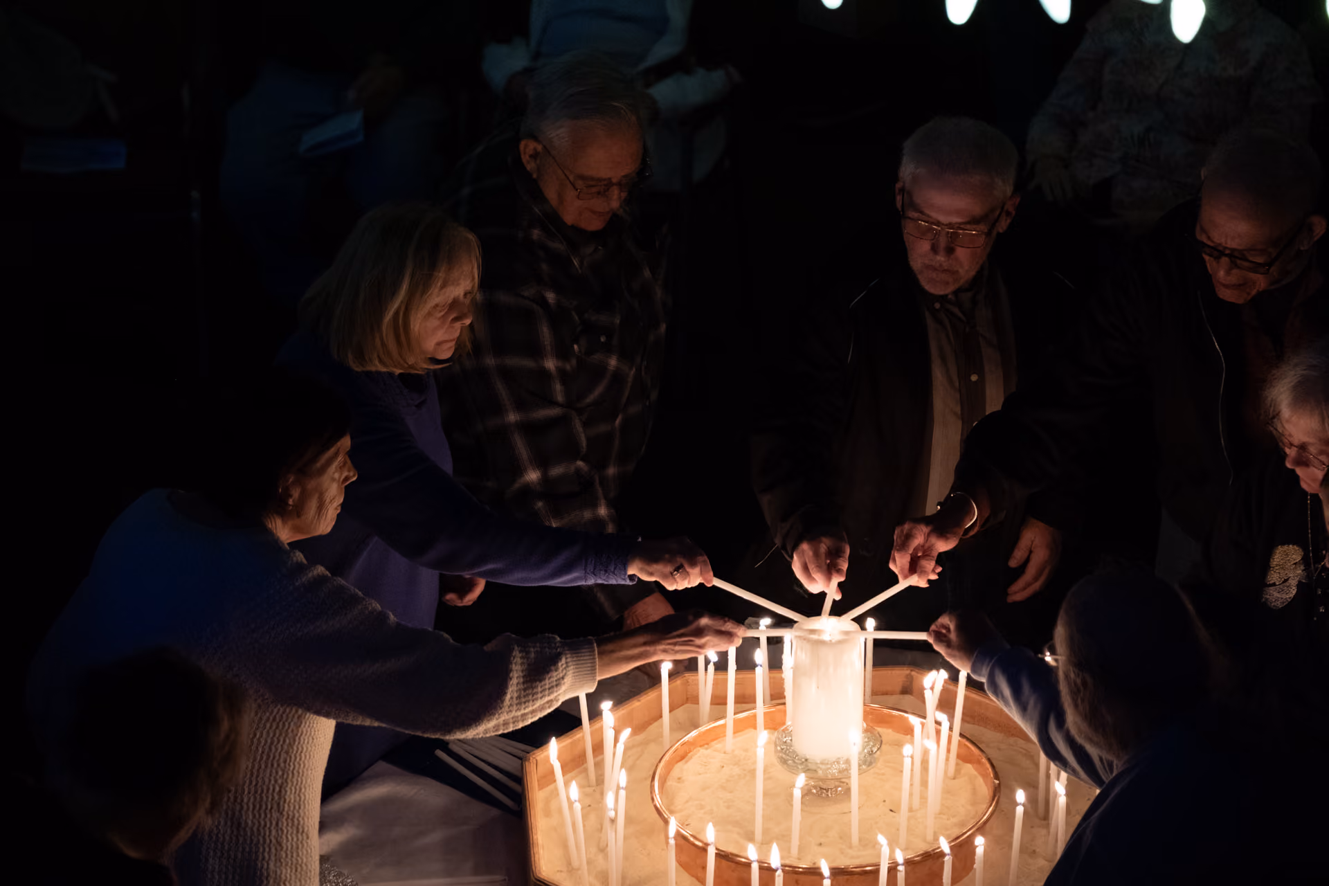 Group of older adults lighting candles from a large central candle in a dimly lit setting, symbolizing a candlelight service or vigil.