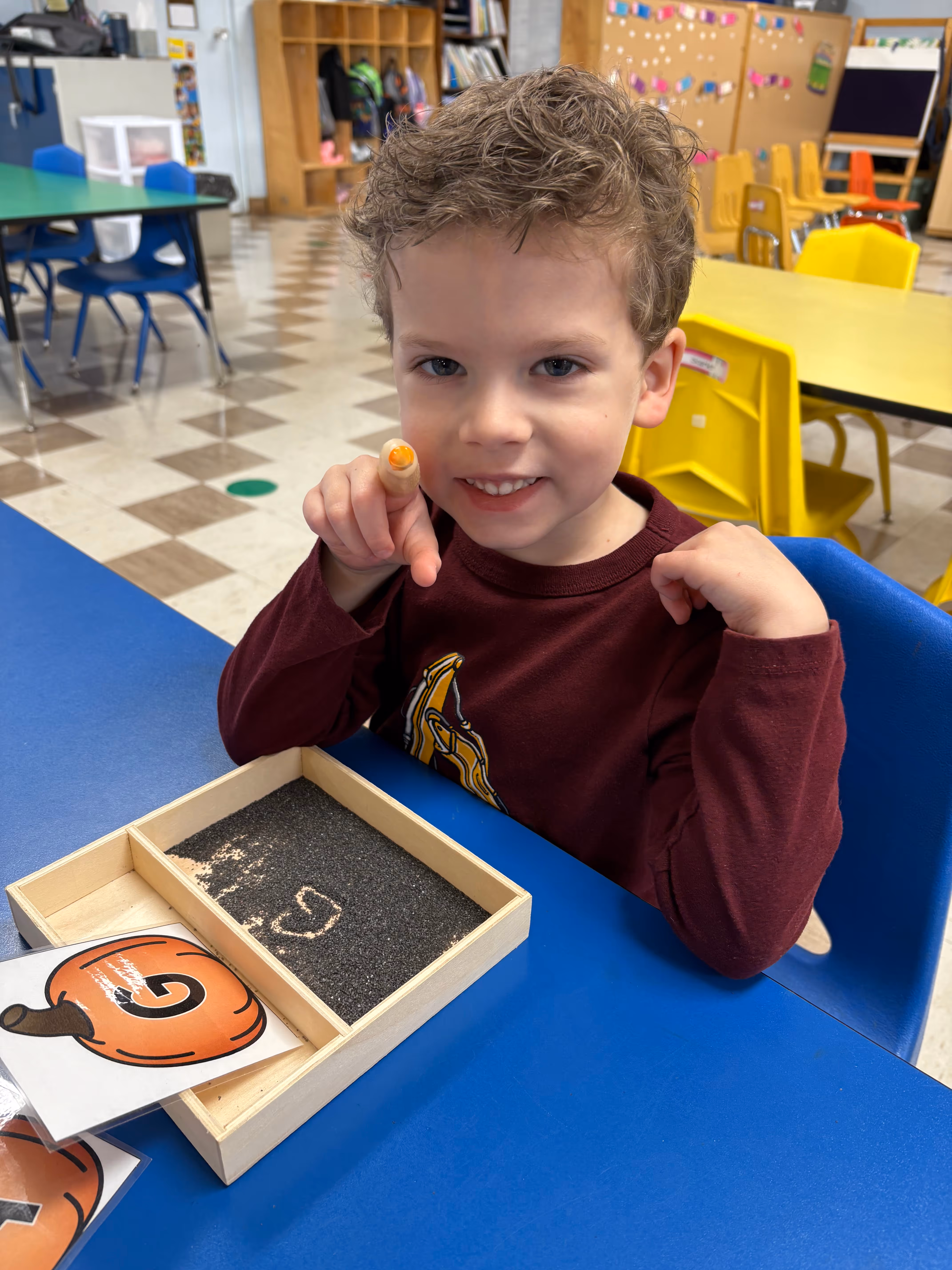 Boy at desk in Good Hope Christian Preschool pointing at the camera and smiling.