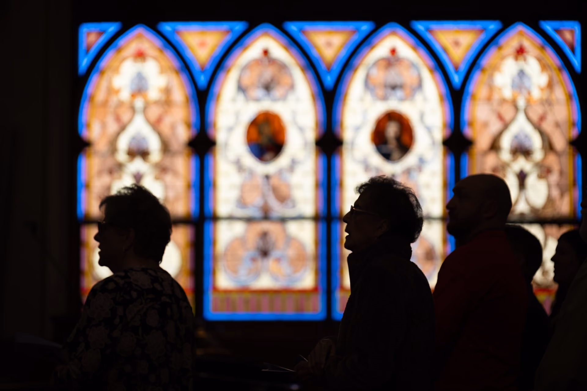 Silhouettes of people standing or sitting inside a church with colorful stained glass windows in the background.