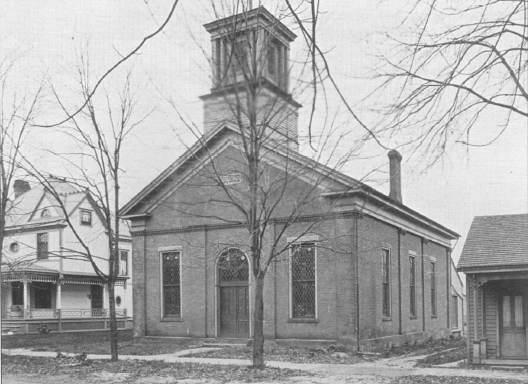 Black-and-white photo of a brick church with tall windows, a central door, a square bell tower, and leafless trees in front.