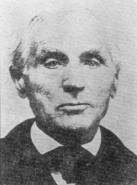 Black and white portrait of an older man with combed-back hair, wearing a dark suit and bow tie, looking straight at the camera.