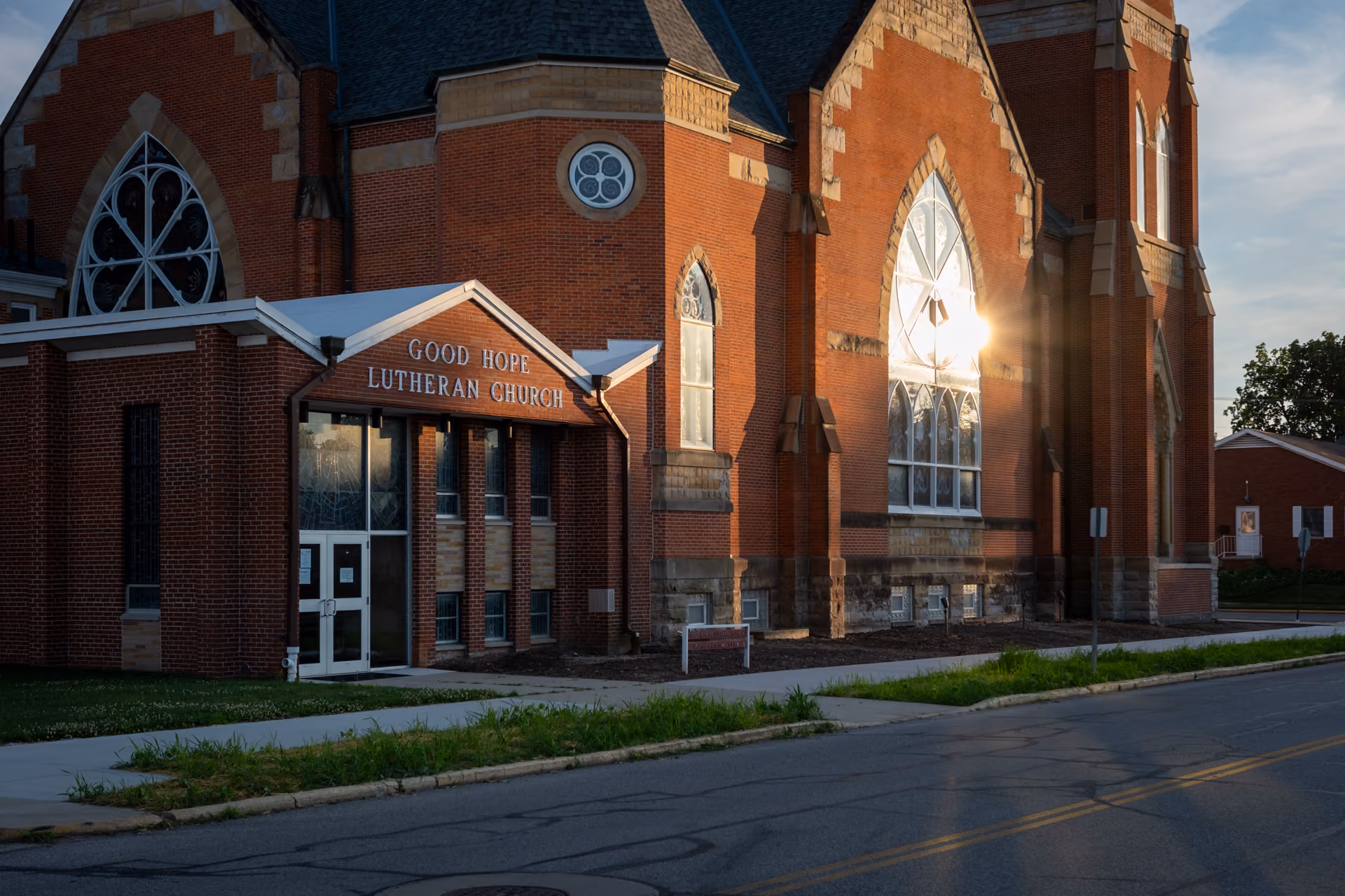 Red brick Good Hope Lutheran Church building with large arched stained glass windows reflecting the sun, a white sidewalk, and a road in front at sunset.