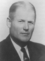 Black and white portrait of a man with slicked-back hair wearing a suit and tie, facing forward with a neutral expression.