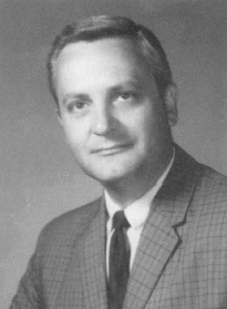 Black and white portrait of a middle-aged man wearing a checkered suit jacket, white shirt, and dark tie, with short neatly combed hair, looking slightly to the side.