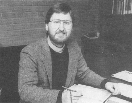 Black and white photo of a man with glasses, beard, and mustache sitting at a desk writing in a notebook, with papers and a brick wall background.