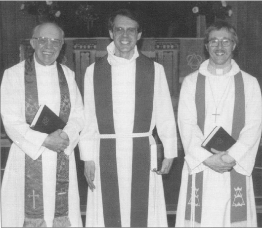 Black and white photo of three male pastors in robes standing indoors, each holding a book and smiling.