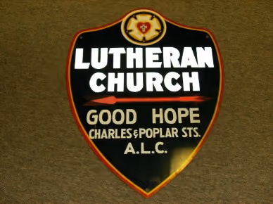 Shield-shaped sign with a red-bordered black background displaying text: Lutheran Church, Good Hope, Charles & Poplar Sts., A.L.C., with a small Luther rose emblem at the top.