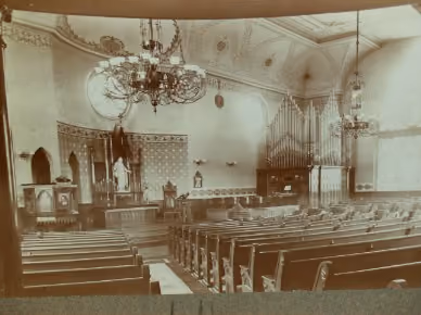 Interior of a vintage church with wooden pews, large chandelier gas lights, ornate organ pipes, tall arched windows, and decorated walls and ceiling.