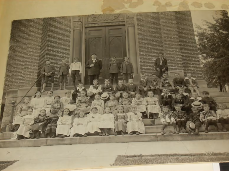 Black and white photo of a large group of children and two adults posed on steps in front of a brick building with large double doors, a portrait from the early 20th century.