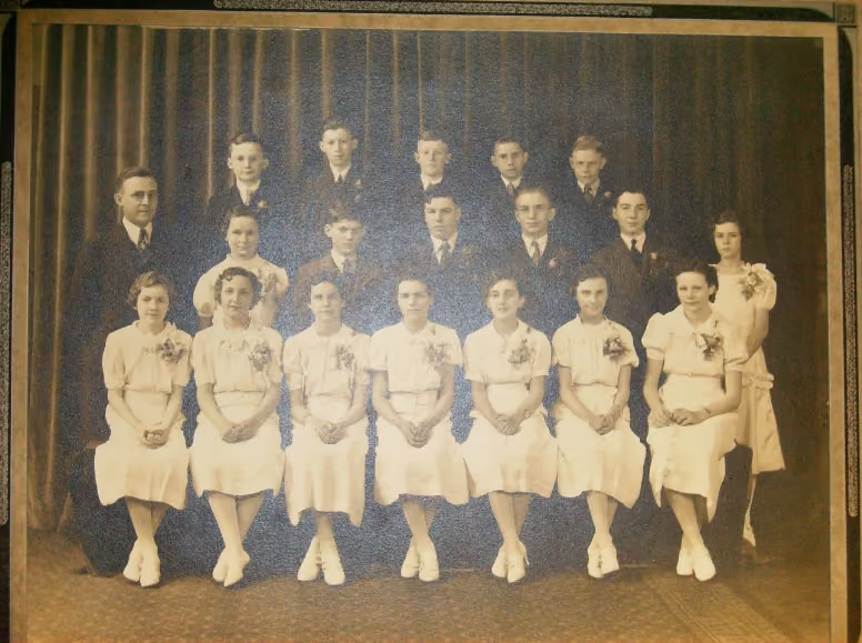 Vintage group portrait of 18 young adults, 11 women in light-colored dresses seated in front row, 7 men in dark suits standing behind them, against a plain dark curtain backdrop.