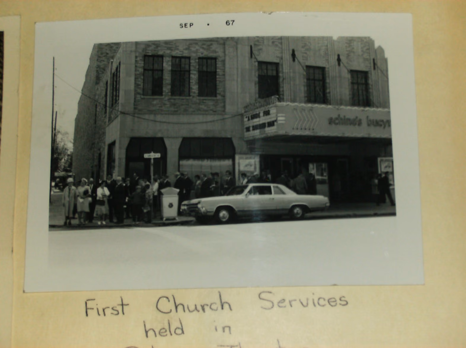 Black and white photo showing a group of people standing outside a theater near a street corner, with an old car parked in front and a marquee sign partially visible.