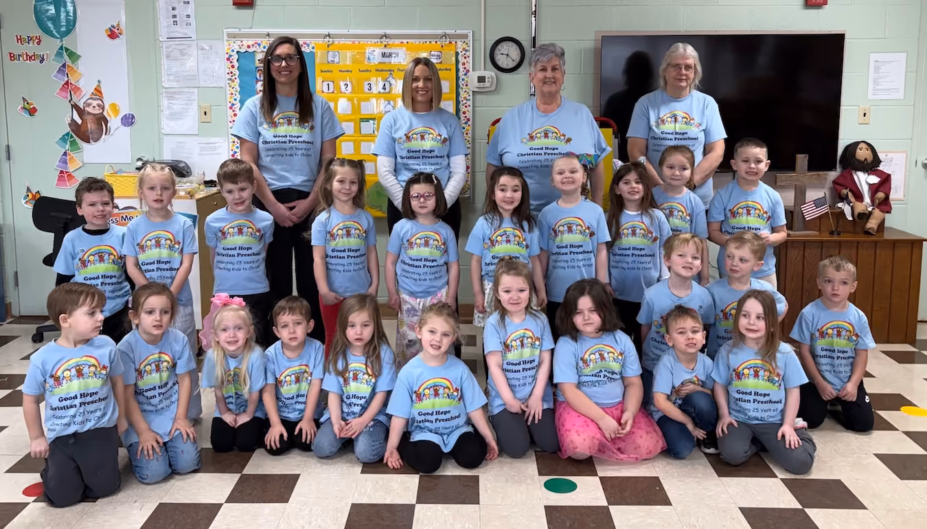 Group photo of preschool children and four teachers wearing matching blue Good Hope Christian Preschool t-shirts in a classroom with colorful decorations and a TV screen in the background.